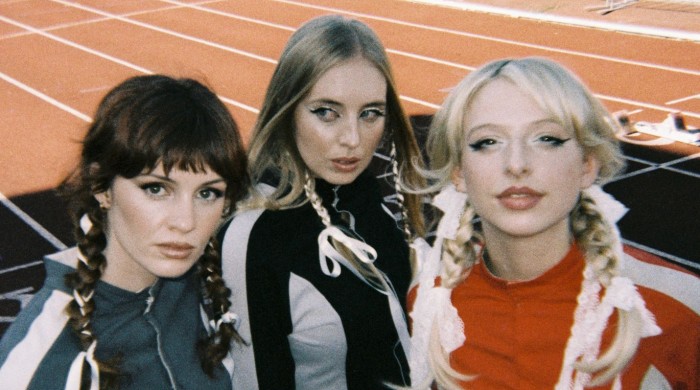 Three members of the band Blusher stood on an athletics track all with plaits and bows in their hair